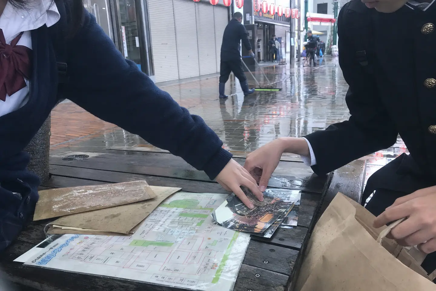 雨上がりの商店街で、二人の生徒が会場図と古い写真を片づける様子
